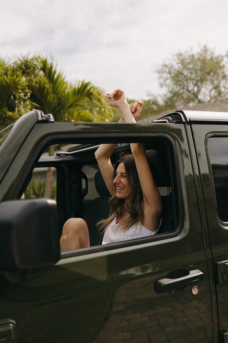 Minnesota and Florida based wedding photographer tosses her arms up in the air with joyful expression on her face as she sits in forest green Jeep.