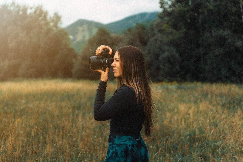 Emily Belanger: Elopement Photographer holding a camera and smiling while standing in a field with a lake in Upstate New York in the background