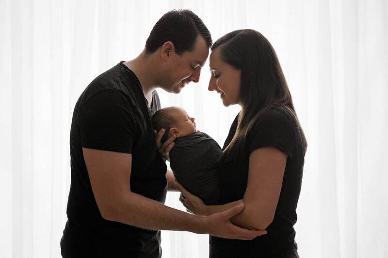 mother and father standing head to head holding their swaddled newborn   baby