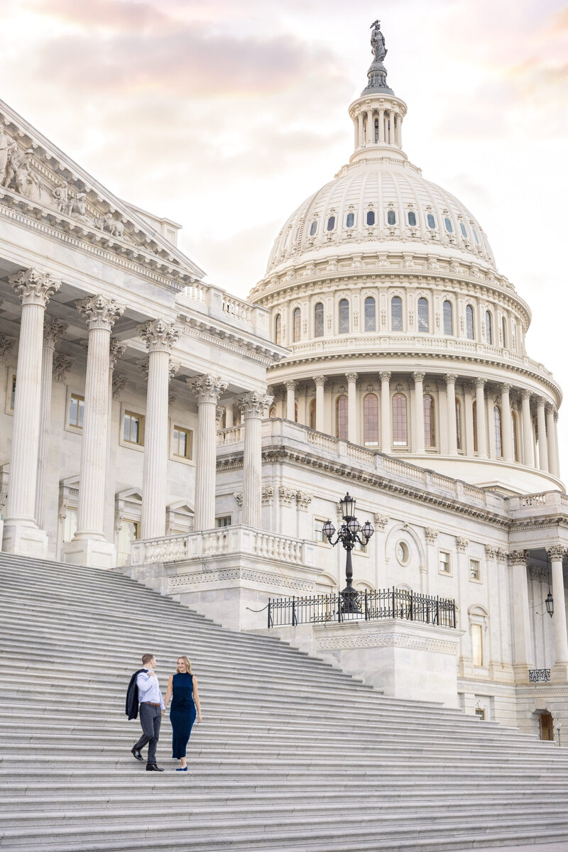 A man and woman walk up the steps of the Capitol, a grand building with large white columns and a dome. The sky is partly cloudy in the background.
