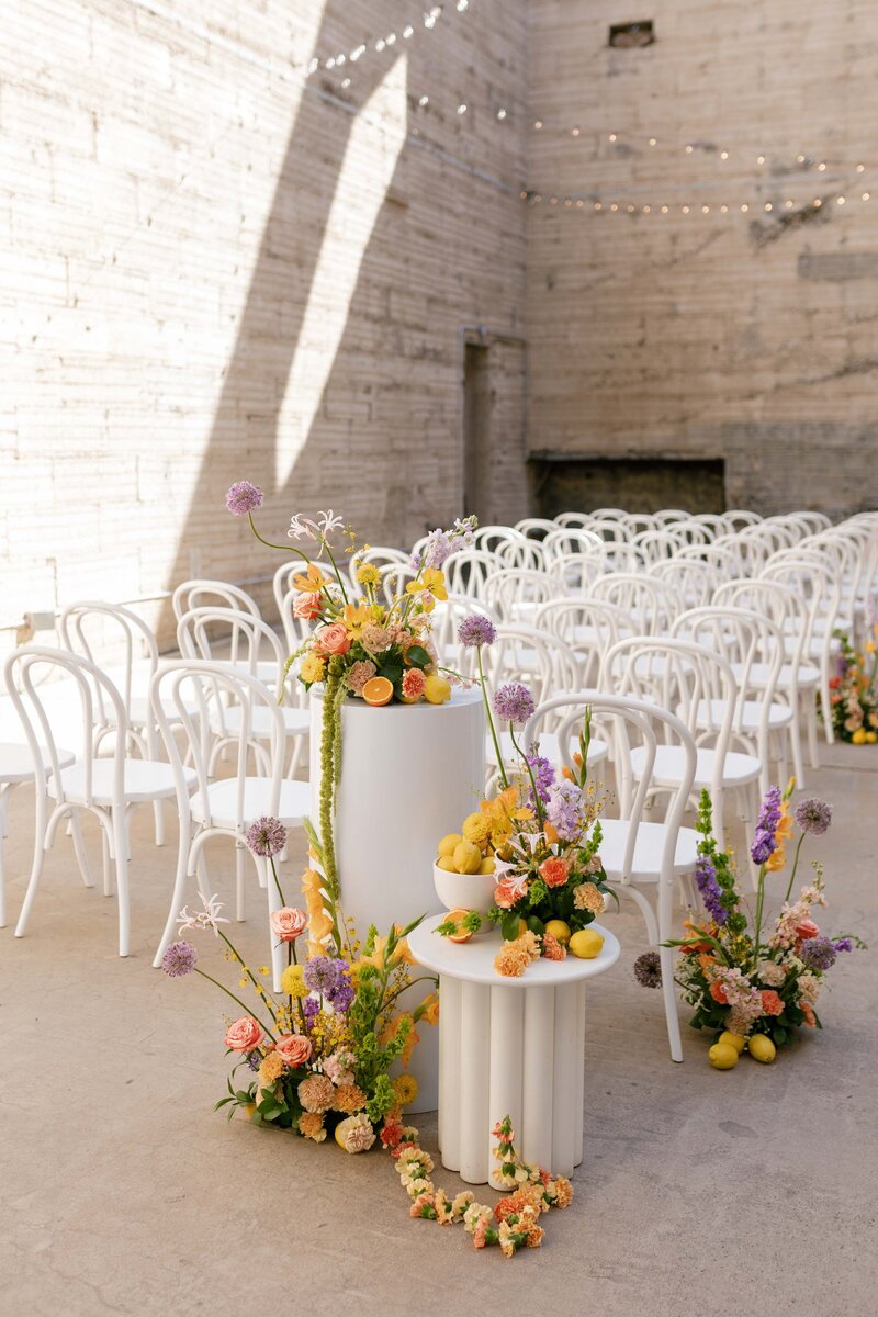Wedding ceremony decor consisting of citrus fruit and colorful florals styled on two-tiered columns.