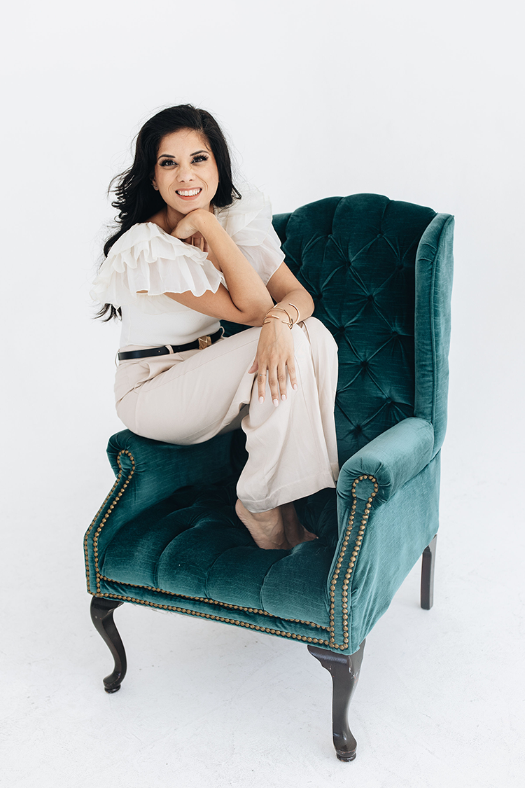 Smiling woman wearing a white top with ruffles and trouser pants, sitting on the arm of a plush teal velvet armchair.