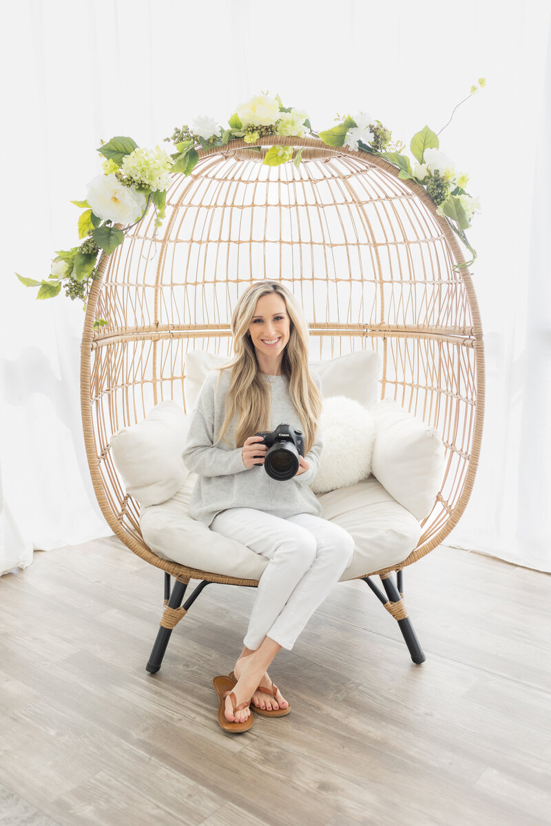 nichole pach sitting on egg chair with floral arrangement holding camera with ankles crossed