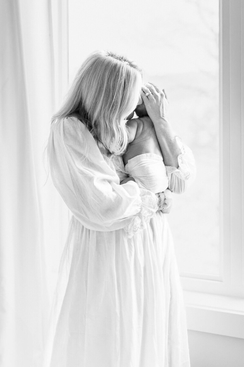 Black and white image of mother in a long white dress gently cradling her newborn while standing by a window during an in-home lifestyle session in Kansas City.