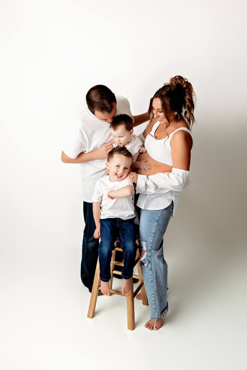 Family of four standing in front of a white backdrop. The mom and dad are tickling the two little boys who are sitting on a stool.