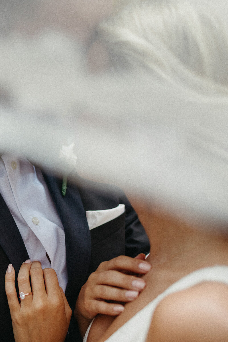 A close-up of a couple in formal attire softly veiled, captured by a destination wedding photographer. The woman, wearing a ring on her finger, gently touches the man's jacket.