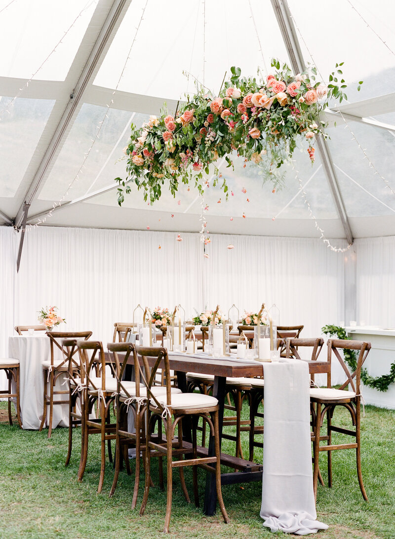 Clear tent wedding reception space. There are long dark brown wooden tables decorated with long white table runners, and pink flowers and foliage hanging from the ceiling above the table.