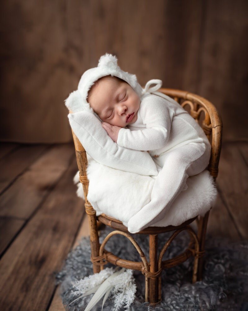 A soft white scene featuring a sleepy newborn boy in a bear bonnet, curled up on a vintage rattan chair.