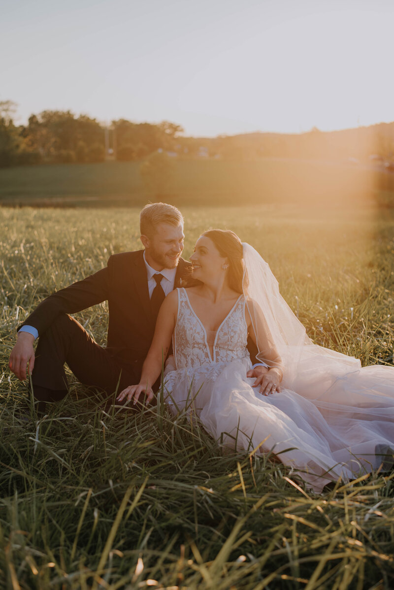 A bride and groom sit together in a grassy field at sunset, embodying the charm of rustic wedding venue photos. The bride wears a white gown with a veil, while the groom is in a dark suit. They smile warmly at each other as golden light surrounds them, capturing their joy.