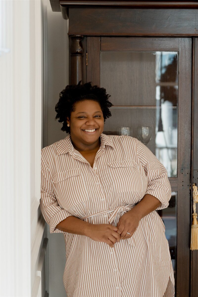 Morgan, founder and creative director of Grey Cove Designs, standing with a warm smile in a neutral-toned striped dress, leaning against a vintage glass cabinet in a sunlit interior.