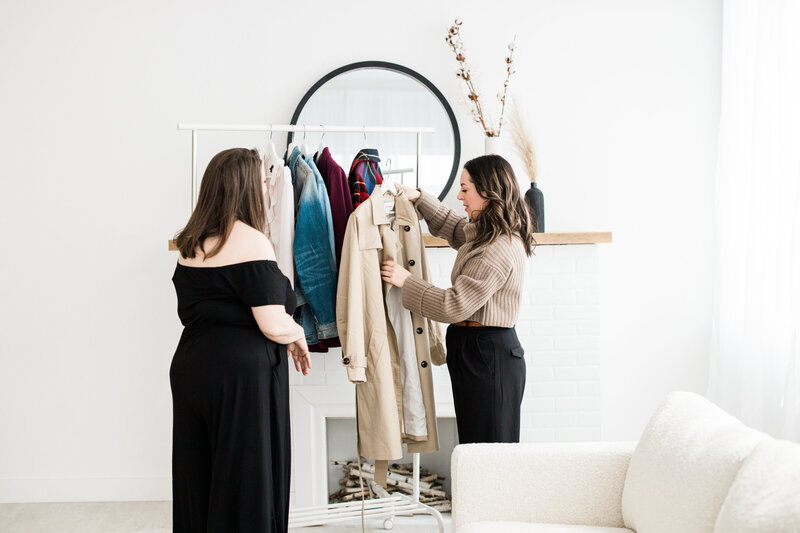 Two women near the clothes rack and picking up a coat to admire.