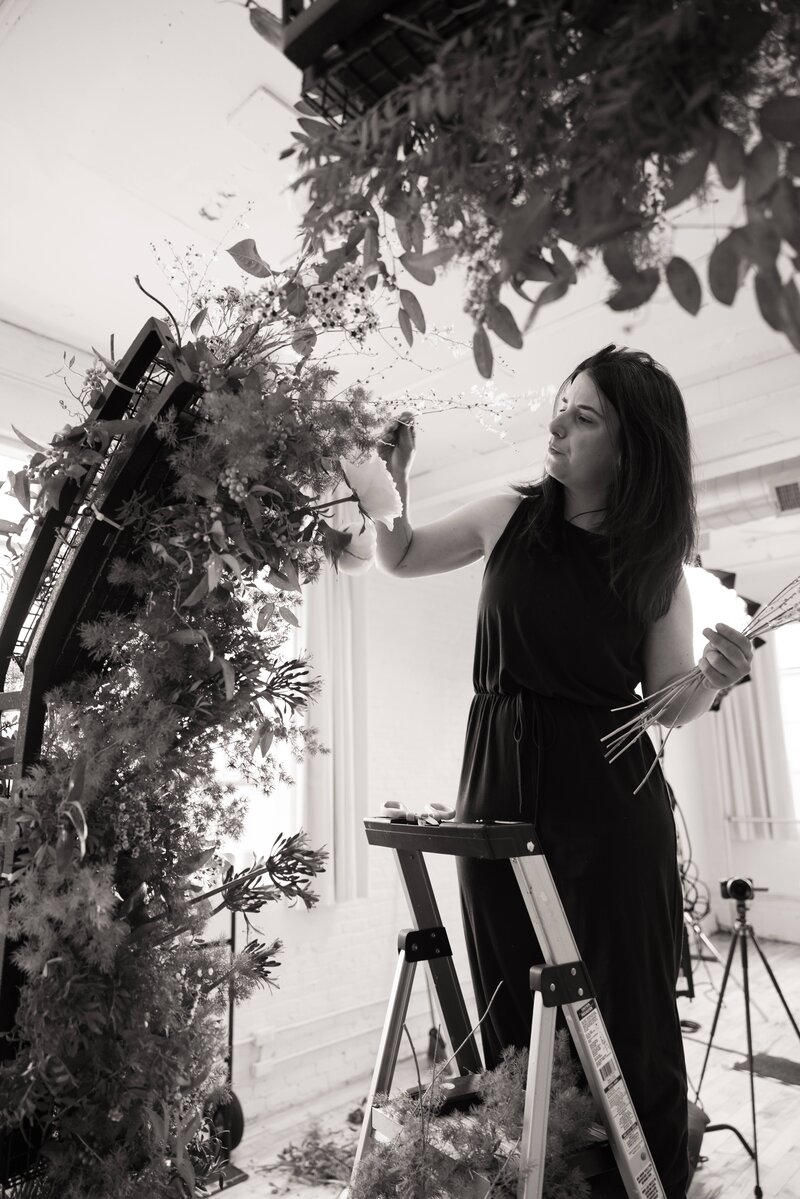 Black-and-white photo of a florist in a sleeveless black jumpsuit reaching up to place flowers into a large, textural floral installation on a standing frame.