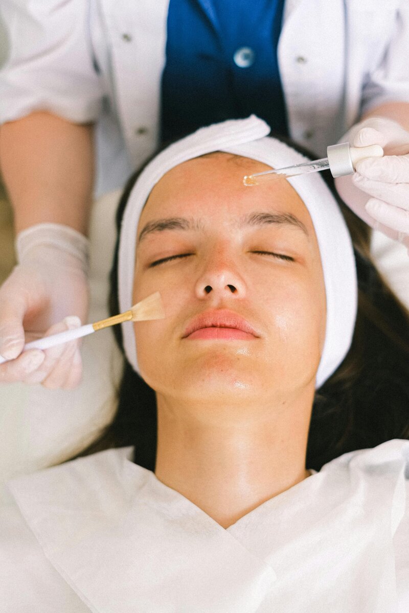 A person receiving a luxurious facial treatment is lying down with eyes closed. An esthetician, in a white lab coat and gloves, gently applies a rejuvenating serum using a dropper and brush. A soft white towel covers the relaxed client, enhancing the serene atmosphere.