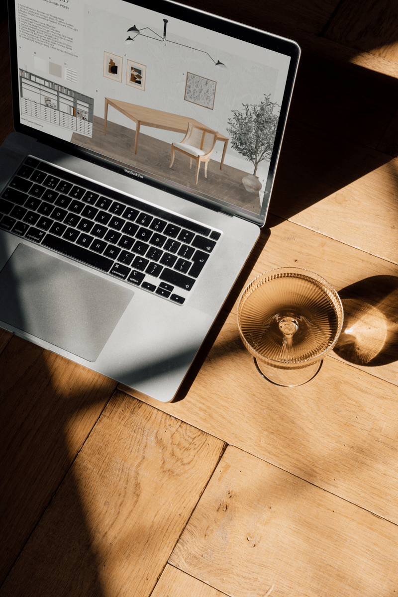Laptop sitting on herringbone timber flooring, with a champagne coupe filled with rosé beside it. Sunlight streams in, casting beautiful light and shadows. The laptop displays a sample board for a formal meeting room designed for an interior design studio.