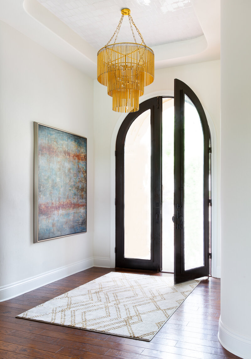 A large arched black framed door with frosted glass opens to a trendy rug with a beautiful gold chandelier overhead in this entryway