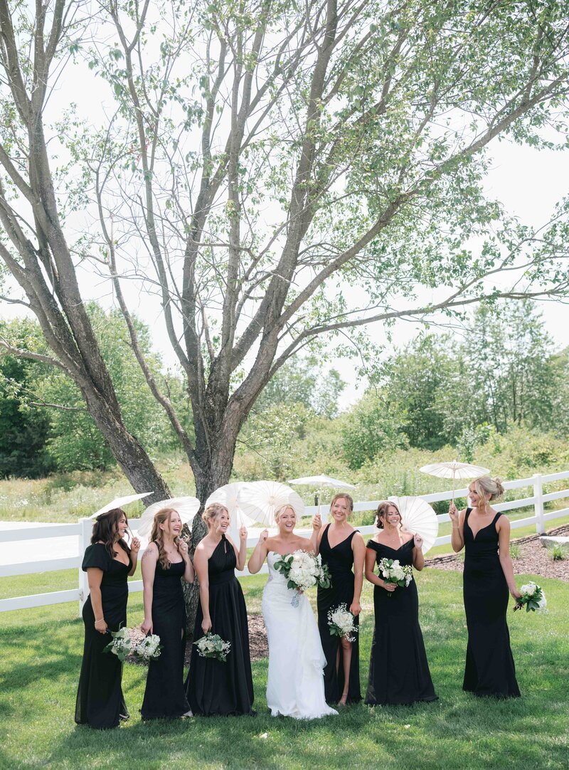 Bridesmaids in black dresses and the bride twirl white parasols as they laugh standing outside on a summer wedding day in Michigan