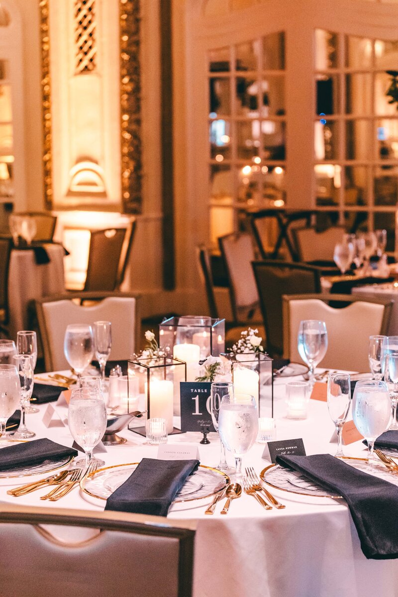 Elegant wedding dining setup in a warmly lit venue with round tables, white linens, and gold cutlery, numbered centerpiece, and candle decor.