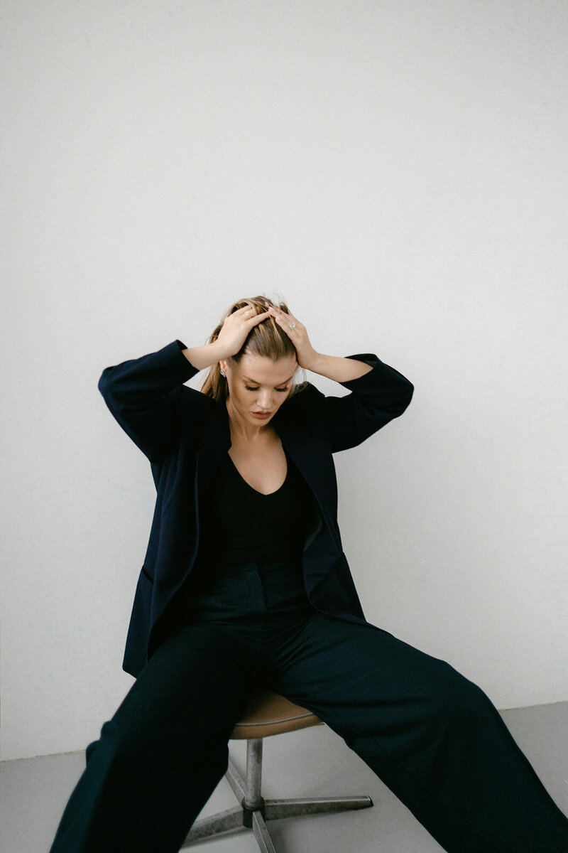 Woman in a black business suit, sitting on a chair, pulling her hair back looking for relief from Financial Stress.