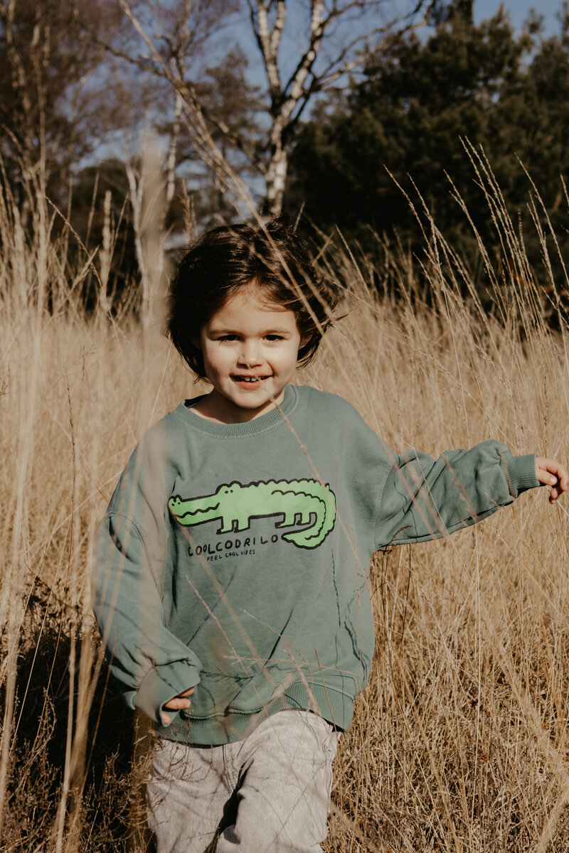 Een jonge jongen in een groene sweatshirt rent door hoog gras, met een blije uitdrukking op zijn gezicht.