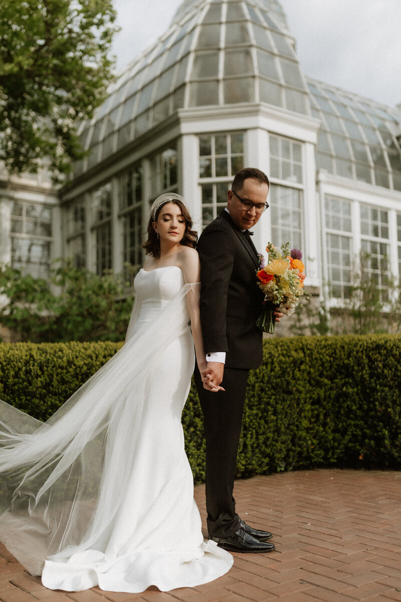 Bride and groom standing hand in hand outside a glasshouse venue, photographed by Daniel from Rainstorm Photo & Video with elegant, editorial composition.