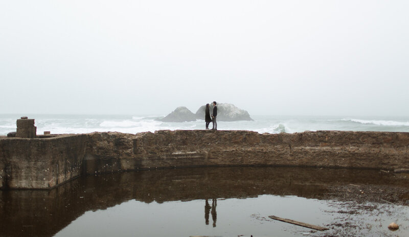 Couple standing on a wall next to the ocean. You can see their reflection in a pool of water in front of the wall.