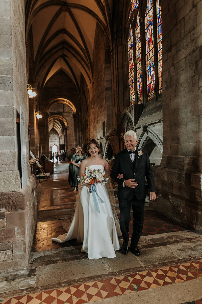 Bride walking to Herefordshire cathedral with her dad holding a bouquet of flowers 