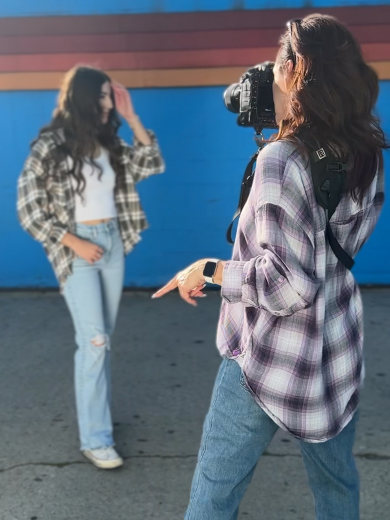 female photographer holding a nikon photographing a high school senior looking down, both wearing plaid shirts and jeans