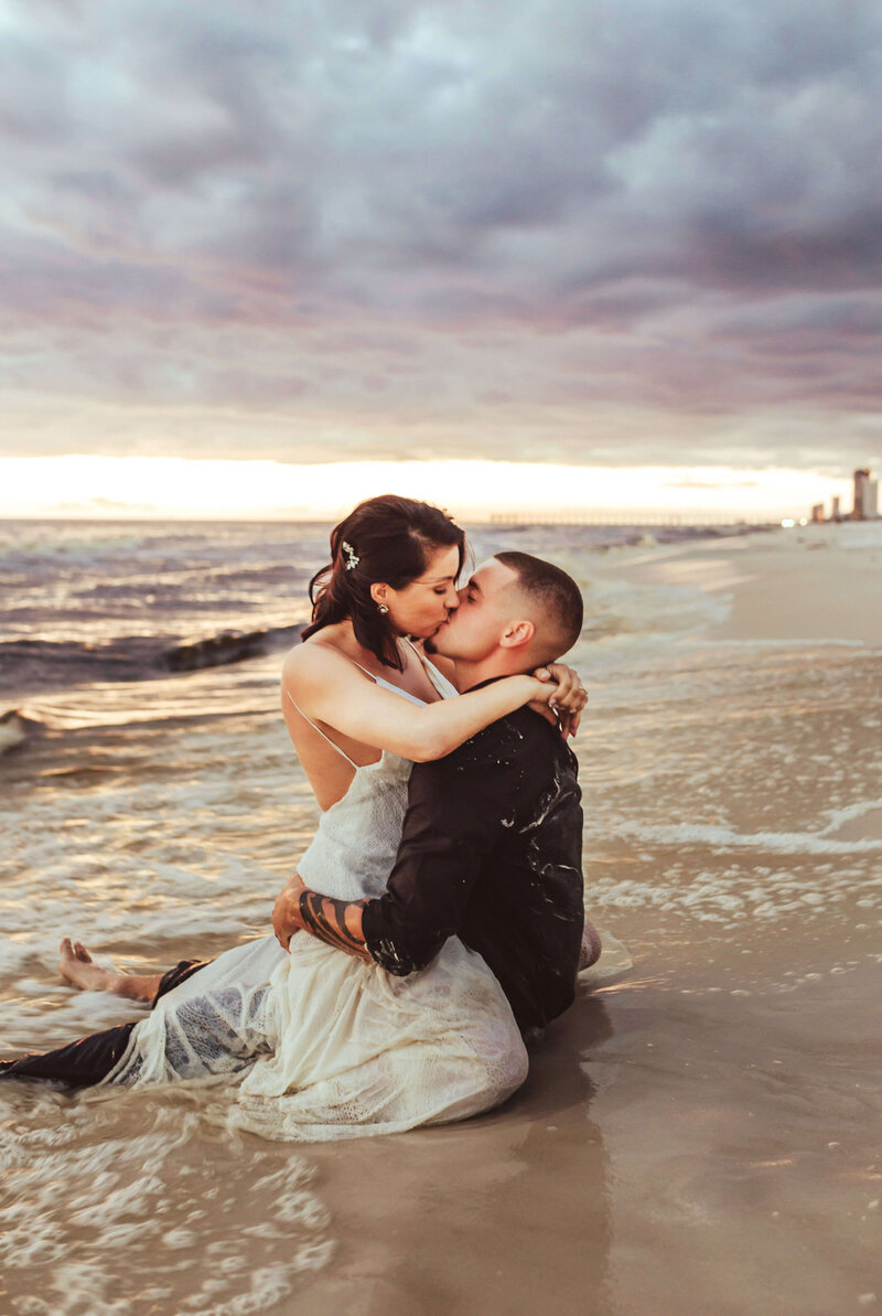 newlyweds sitting on beach in water, kissing at sunset in PCB florida