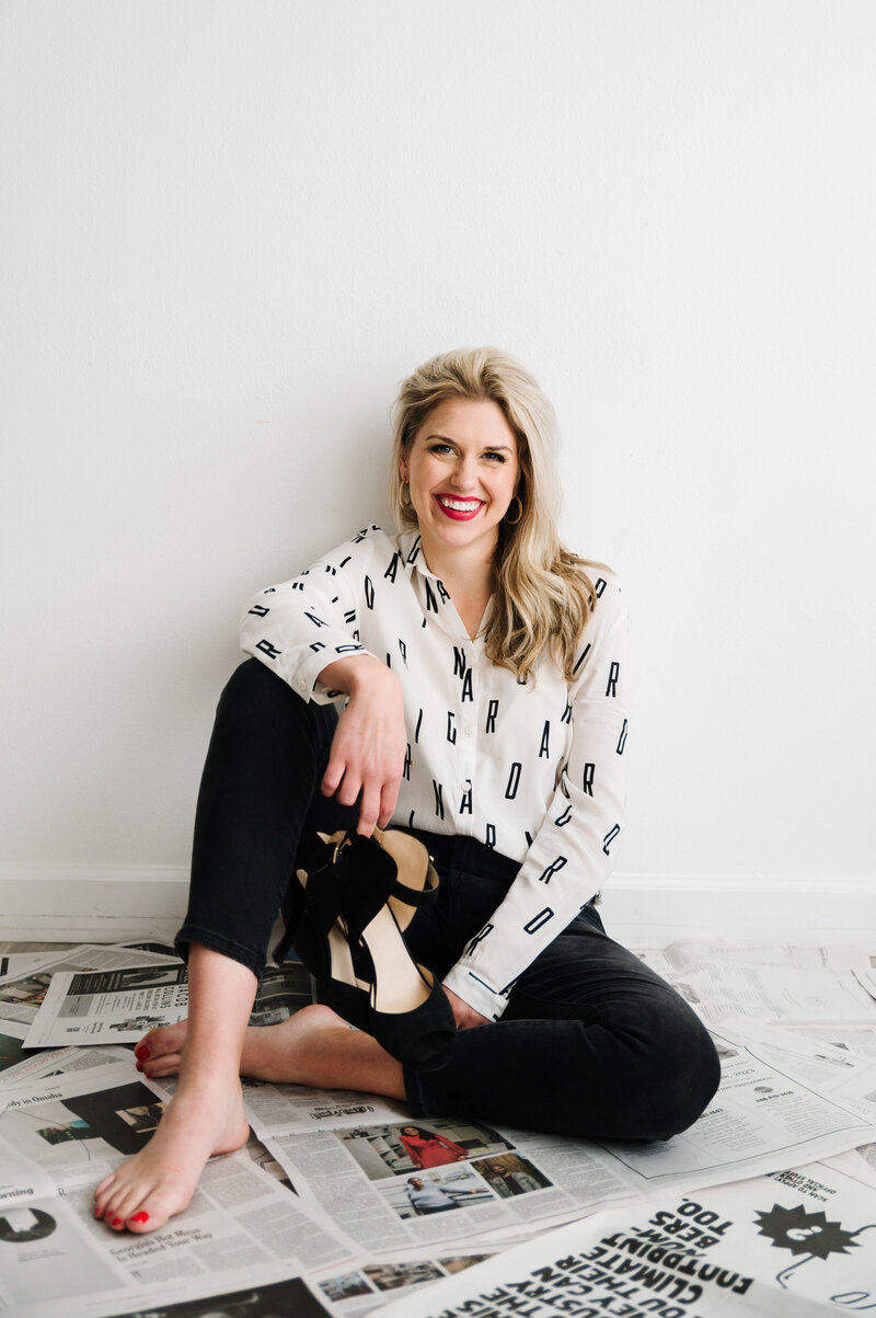 Sarah Klongerbo standing in front of magazine pages hanging on clothespins and wearing black jeans and a white top with letters all over it