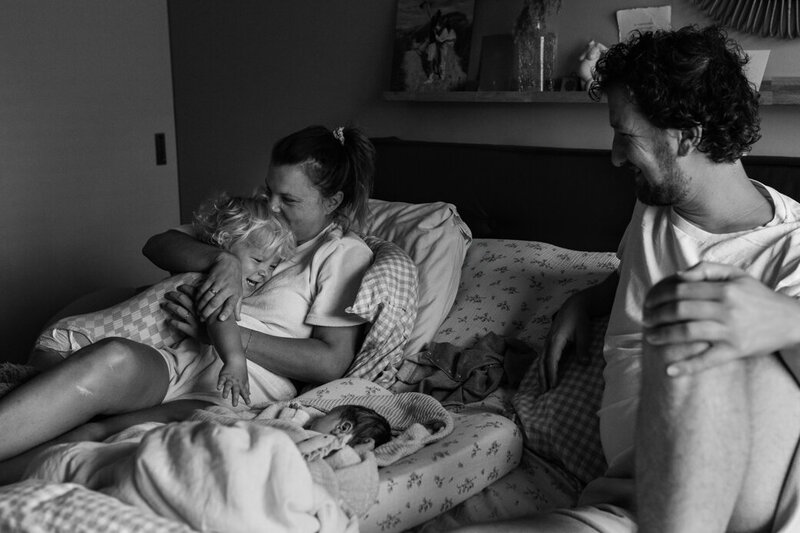 Gezin op bed, baby in het midden. Mama knuffelt grote broer. Papa kijkt liefdevol toe. 