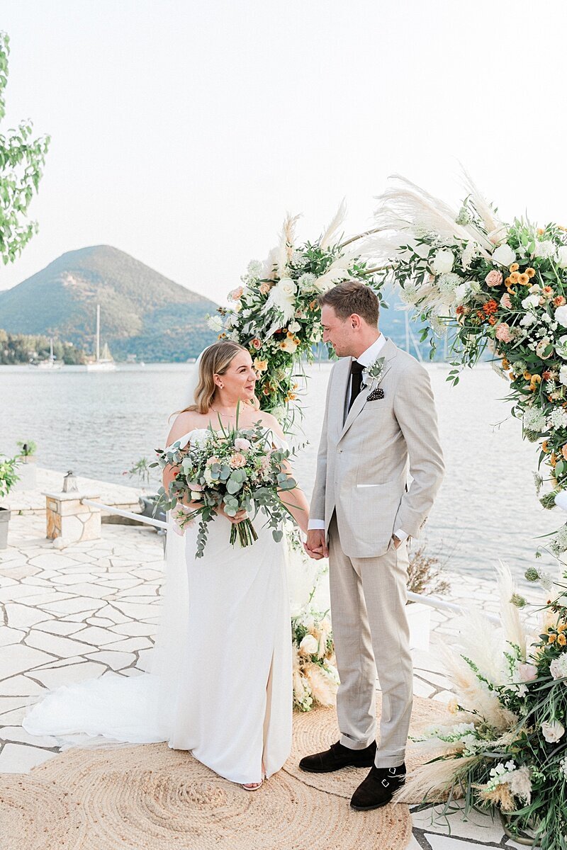 Newlywed couple holding hands in front of a pampas grass and rose floral arch, with coastal mountains and sailboats in the background during their destination wedding in Greece