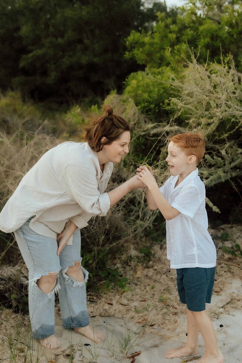 A woman and a young boy stand outdoors near greenery, facing each other and smiling as the boy shows her something in his hands captured by a Middle GA Photographer