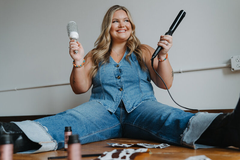 A young lady is sitting on the floor in a studio holding a brush and curling iron. There are hair coloring items in the floor in front of her. She is wearing a denim outfit and smiling off to the side. Branding photos for hair stylist. 