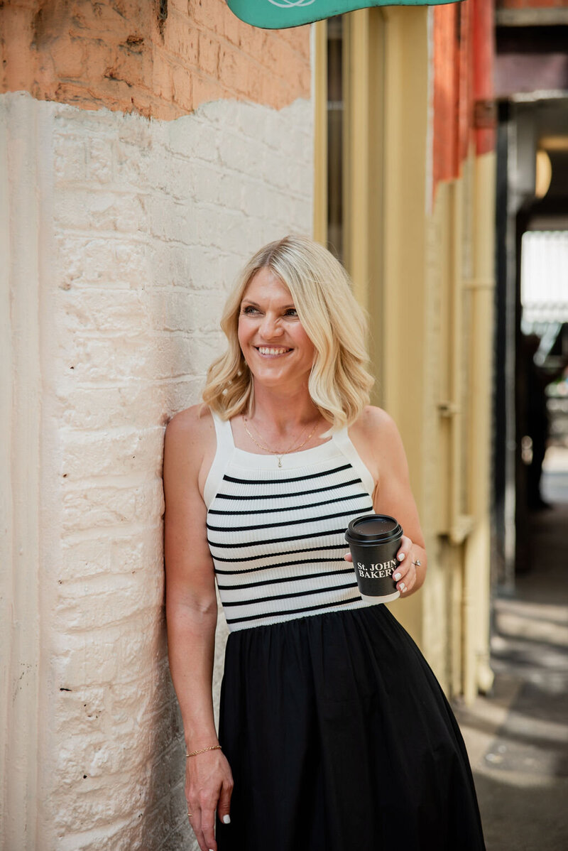 Emily Collins smiling and holding a coffee cup while leaning against a brick wall, with “Hello Lovely” in script above her