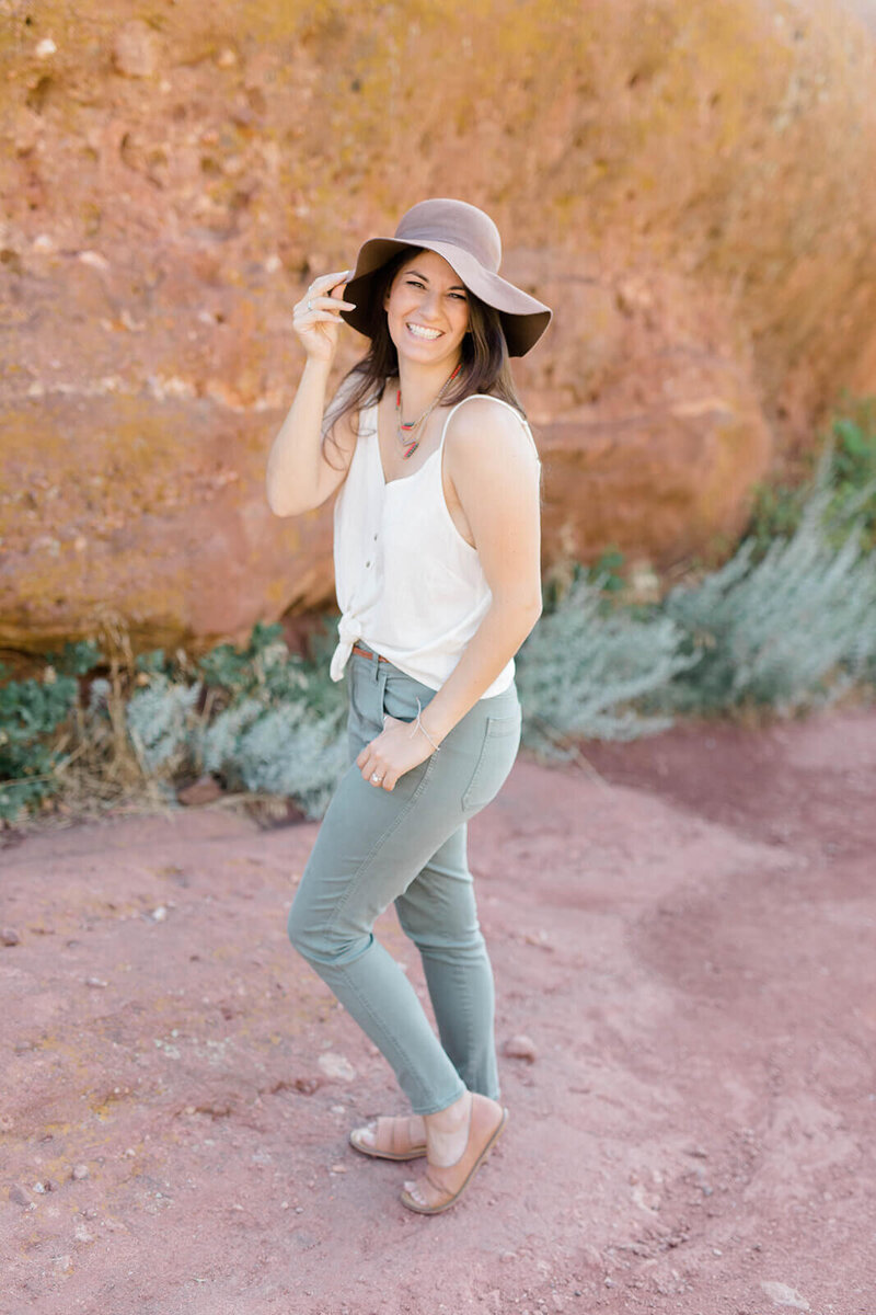 Founder and business strategist of Find Your Freedom Co., Laura. She is standing in front of a large red rock formation wearing green jeans, a white tank and a brown hat.