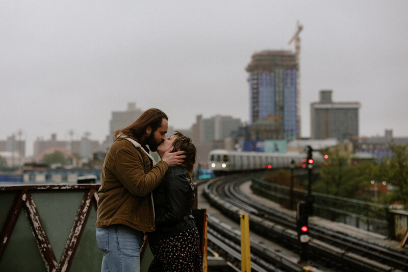 In NYC, a couple kisses on a platform as a train approaches on the tracks nearby, capturing a romantic moment.