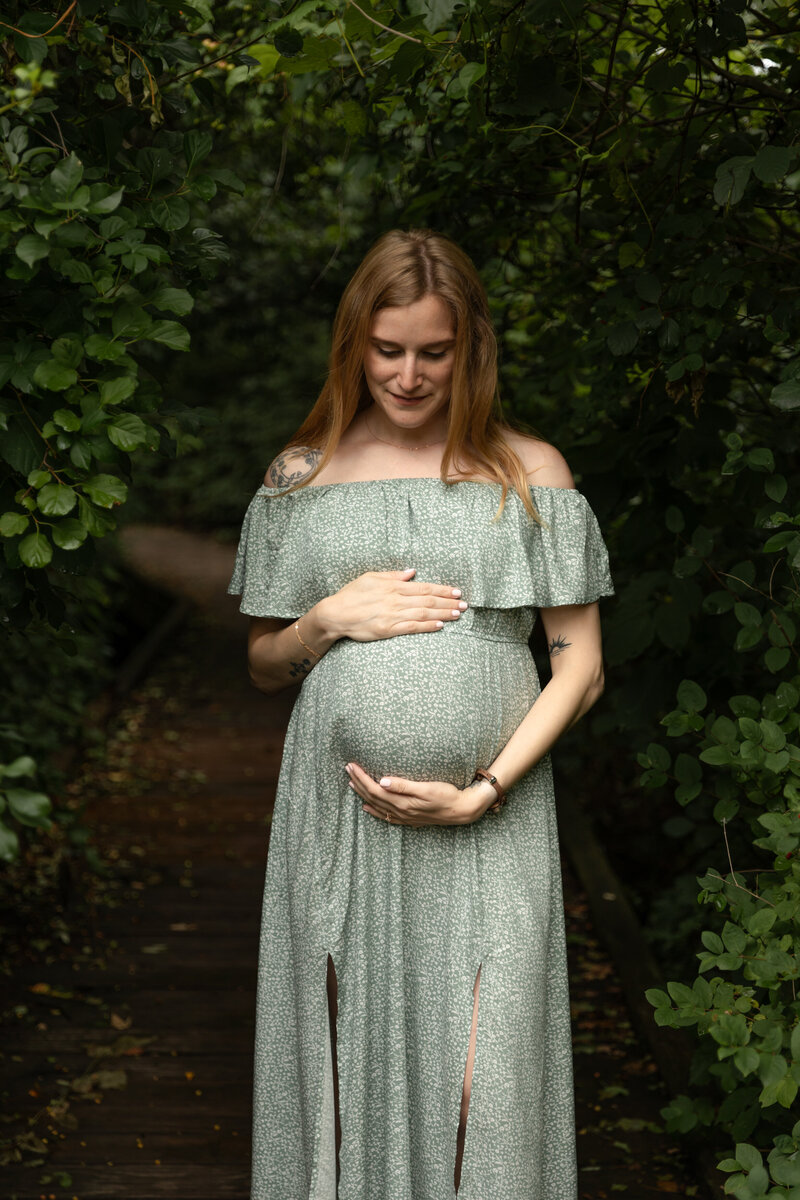 expecting mother looks down at her belly and cradles it on a boardwalk in the woods at Saginaw Forest in Ann Arbor michigan