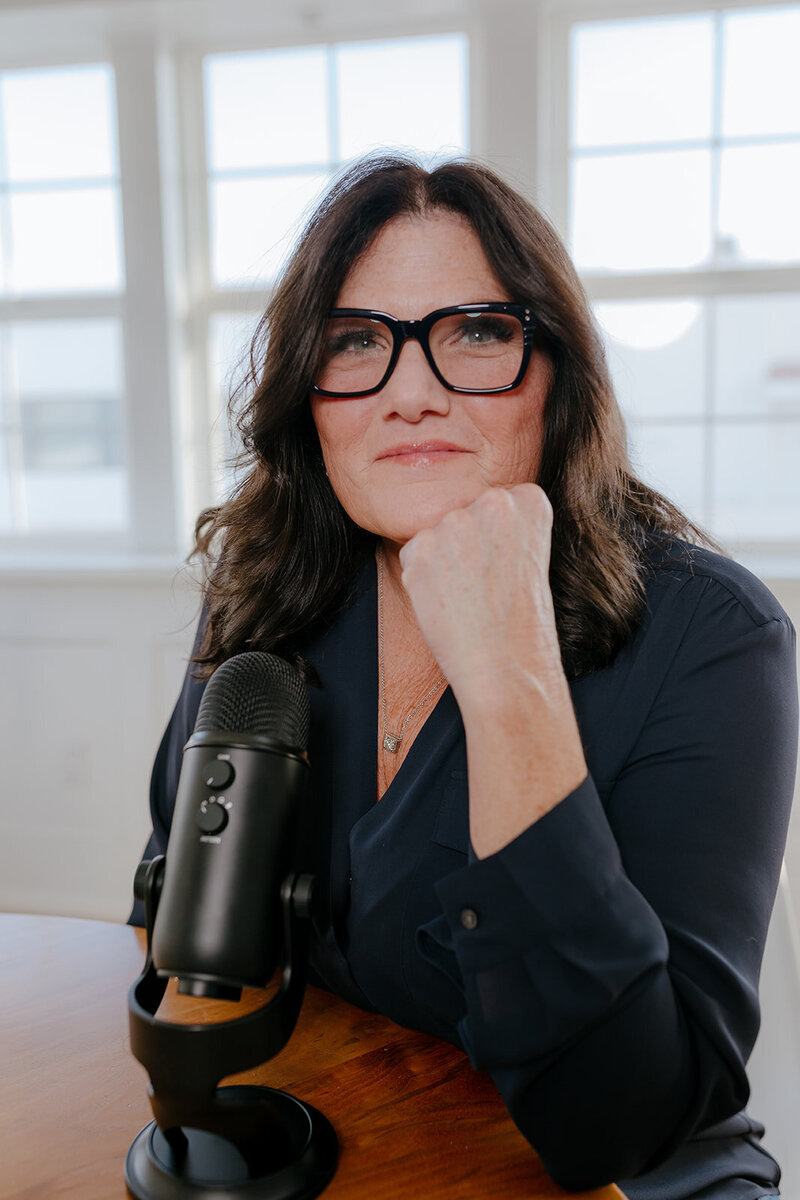 Close-up of Susie Schumacher wearing glasses and a navy blouse, seated at a wooden table with a podcast microphone, looking confidently at the camera - Susie Schumacher Life Coach