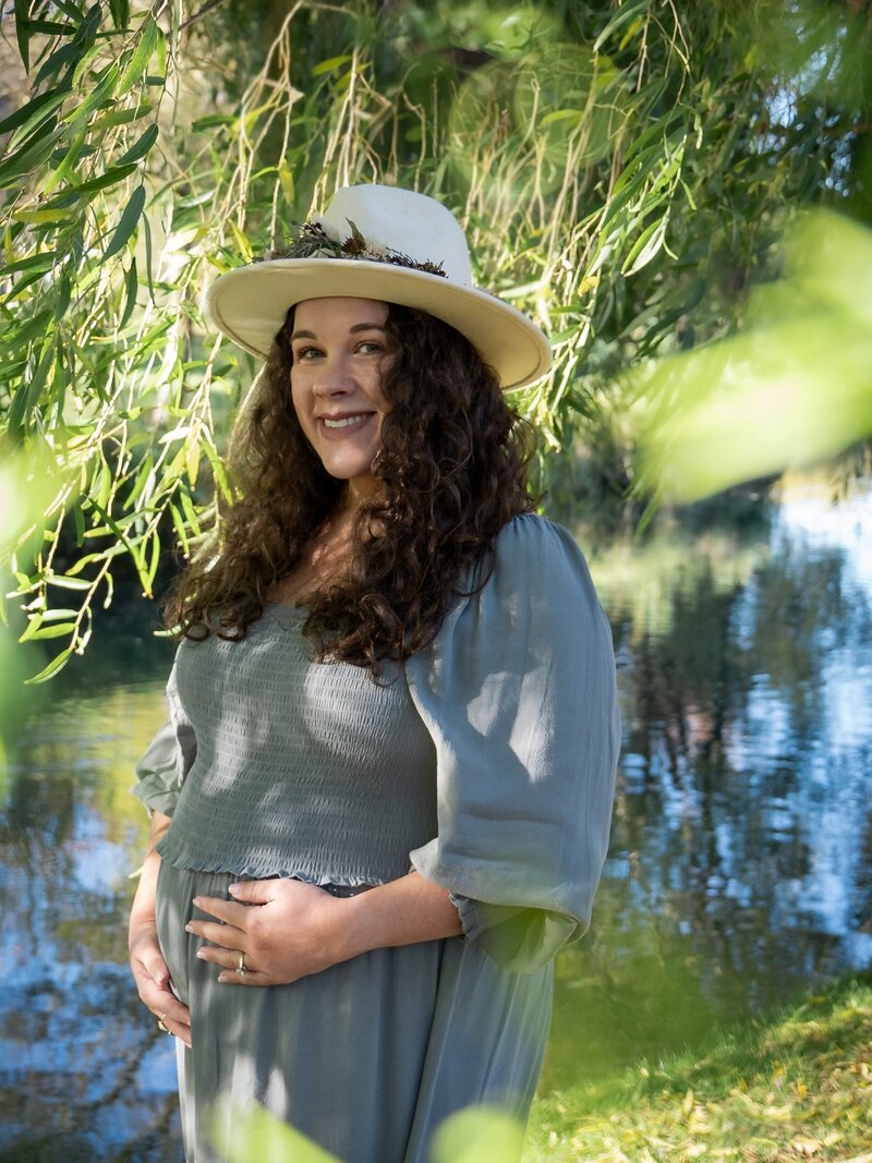 summer maternity session with a pregnant woman. she has dark curly hair and wears a hat and green dress. she is at a park in Christchurch, New Zealand, and there are willow branches surrounding her and a river in the background.