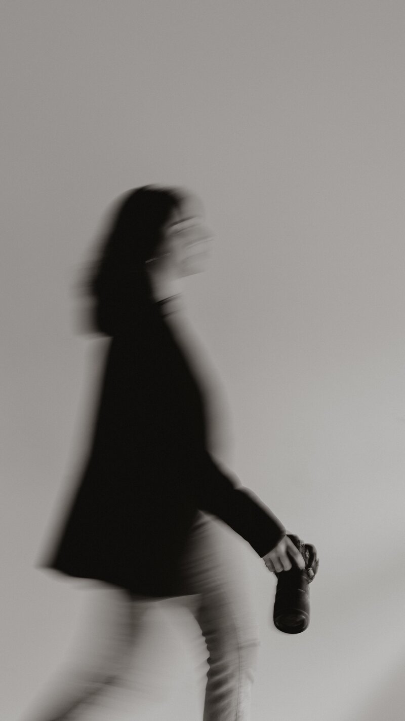 A blurred image captures a person walking with a camera in their right hand, embodying the subtle artistry of a New England portrait photographer. Clad in a dark jacket and light pants, they move against a plain, neutral background, evoking timeless storytelling through every stride.