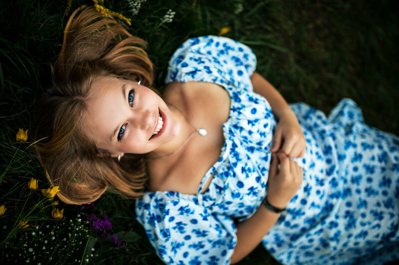 a dynamic portrait of a senior in high school lays in the grass with bright flowers around her and on her dress.