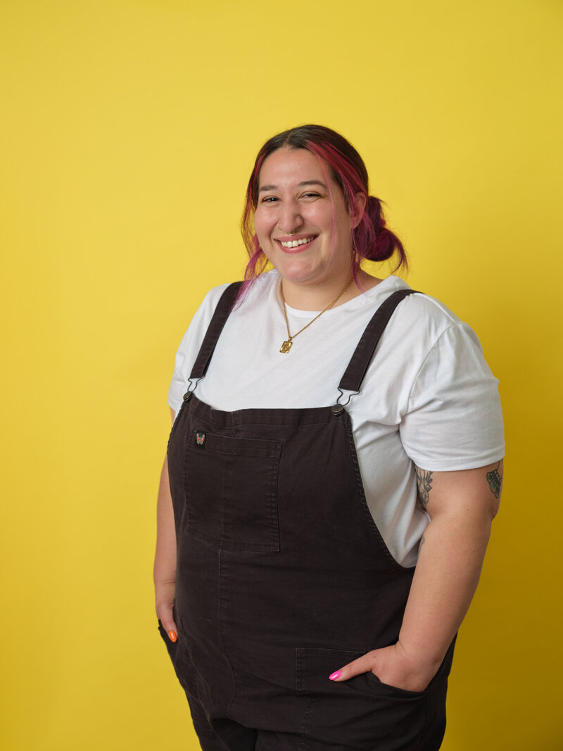 A person in overalls smiling with their hands in their pocket
