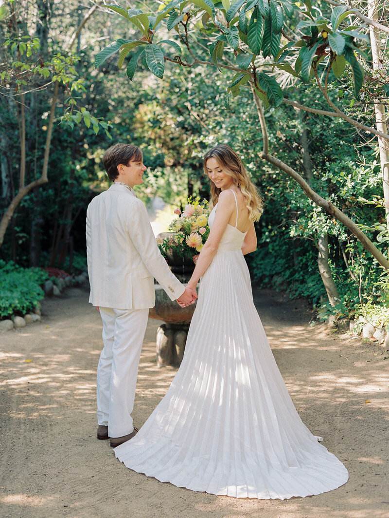 groom looking at bride wearing a white silk pleated gown in a garden