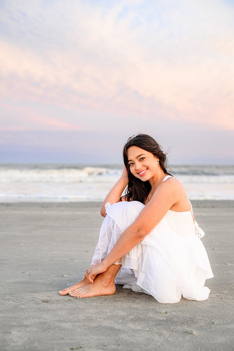 senior girl from waxhaw, nc with long dark hair sits on a charleston, sc area beach wearing a white dress during her destination graduation senior photos.