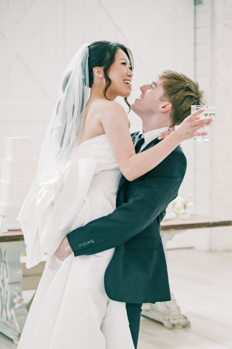 The Nest at Ruth Farms Wedding Venue | DFW Wedding Photographer Rae De Lain Light and Airy | Groom carries bride holding a champagne glass