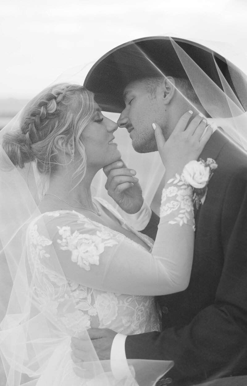 Black and white photo of a groom in a cowboy hat and a bride about to kiss, her veil draped over both of them.