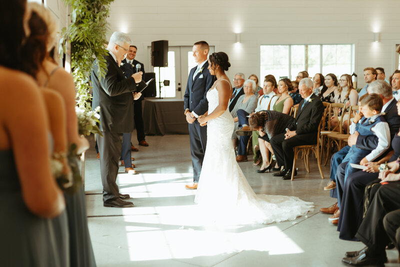 Couple is getting married in the summer at The Barn at Grace Hill. The officiant is leading the ceremony as guests watch expectantly for them to read their vows,