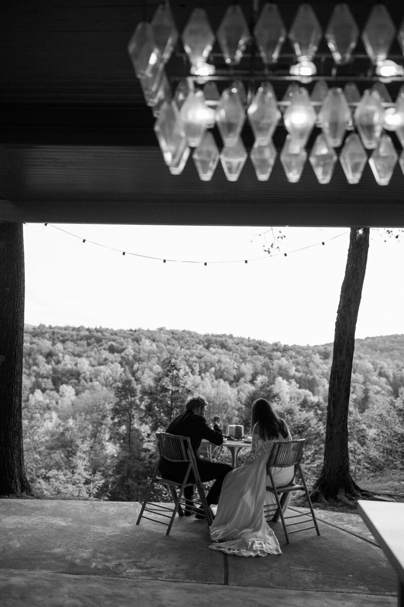 A couple sitting together at a table, overlooking a scenic forest view, with a rustic chandelier hanging above. Black and white photo.