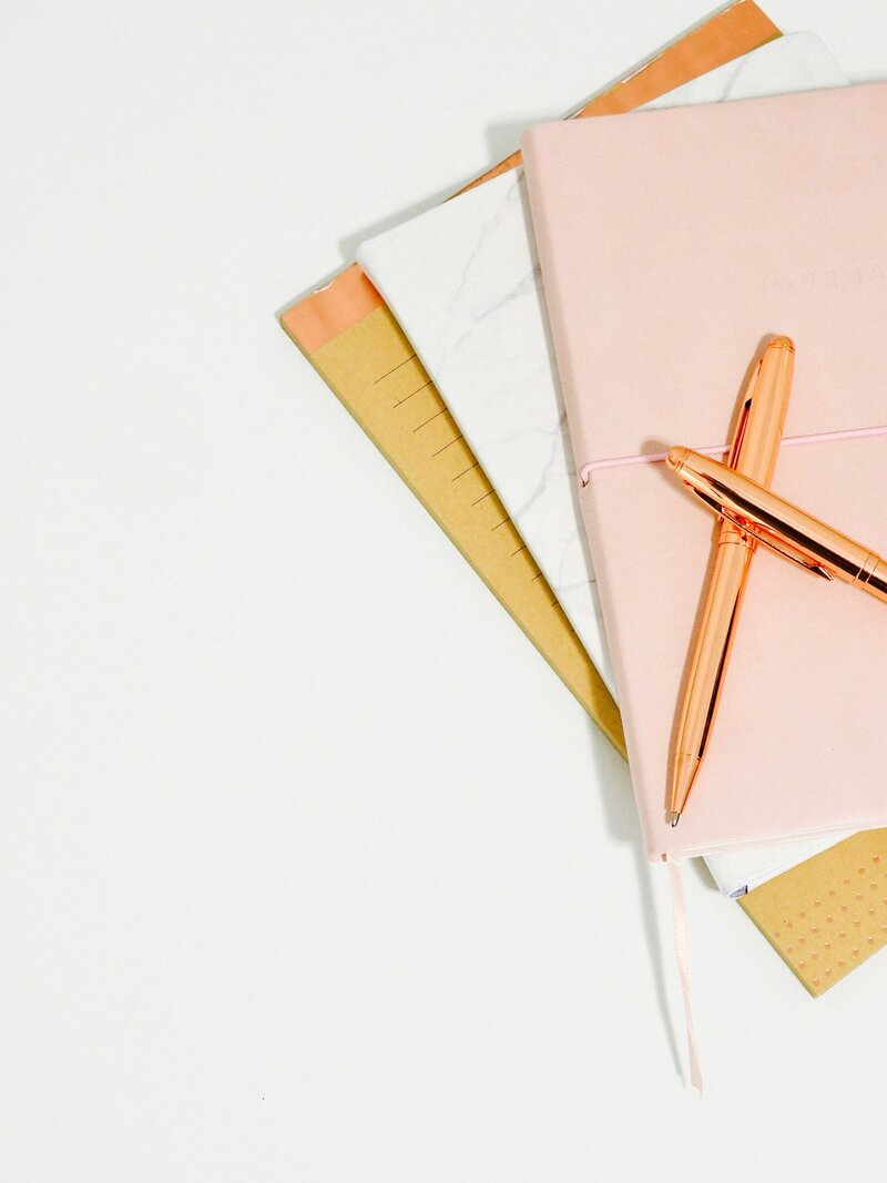 Stack of pink, yellow and white notebooks on a white background with rose gold pens on top