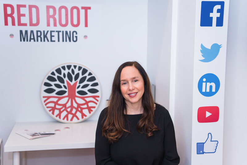A woman sitting at the Red Root Marketing office in Verona, New Jersey with the business logo and social media icons in the background.